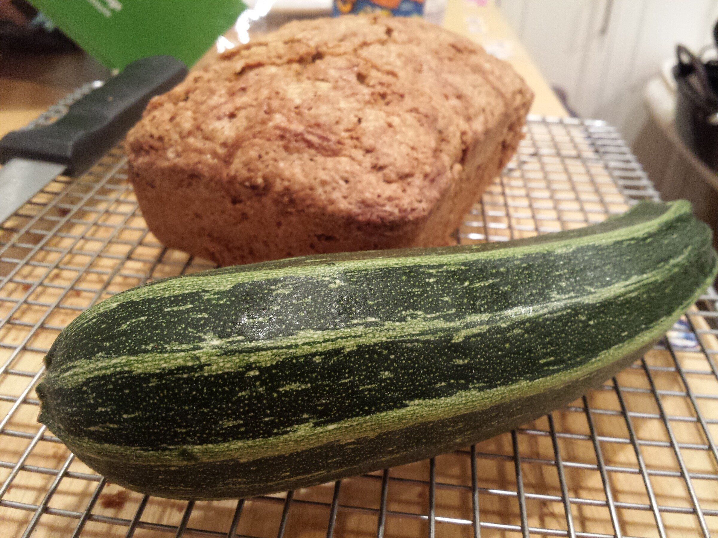 A loaf of zucchini bread on a cooling rack behind a large striped zucchini